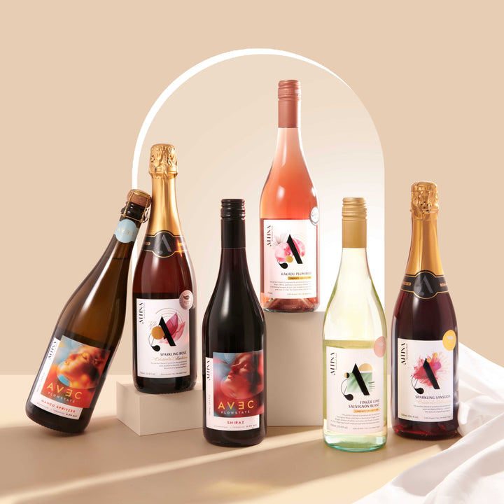 Altina Discovery Pack featuring six bottles arranged against a warm beige background. Two bottles are elevated on plinths: Sparkling Rosé on a lower block and Kakadu Plum Rosé on a taller block. The remaining four bottles stand on the surface, with the Mango Spritzer leaning against the Sparkling Rosé. A soft white fabric is on the right and a subtle arched shape is visible in the background.