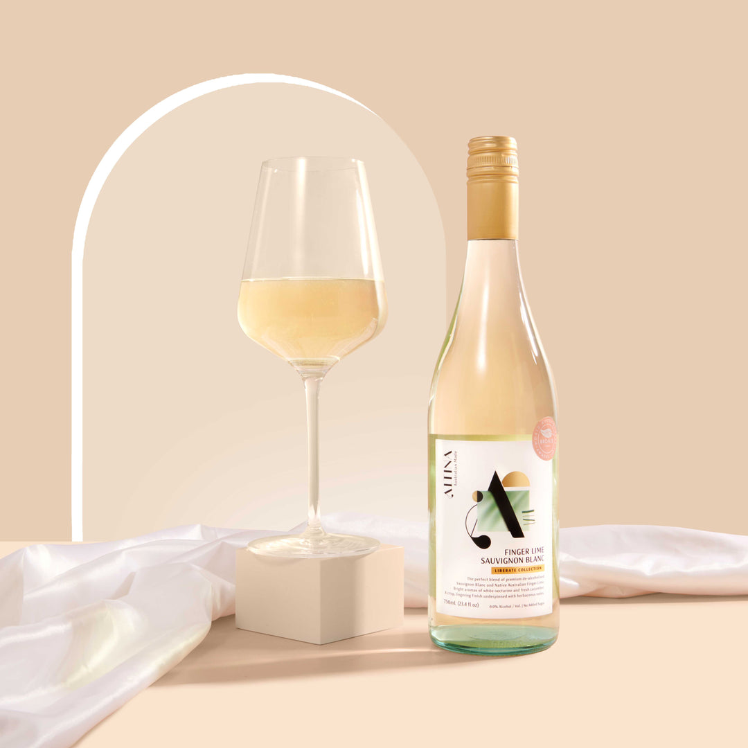 A glass of pale white wine beside an Altina Finger Lime Sauvignon Blanc bottle on a beige surface, with soft white fabric and a curved arch backdrop