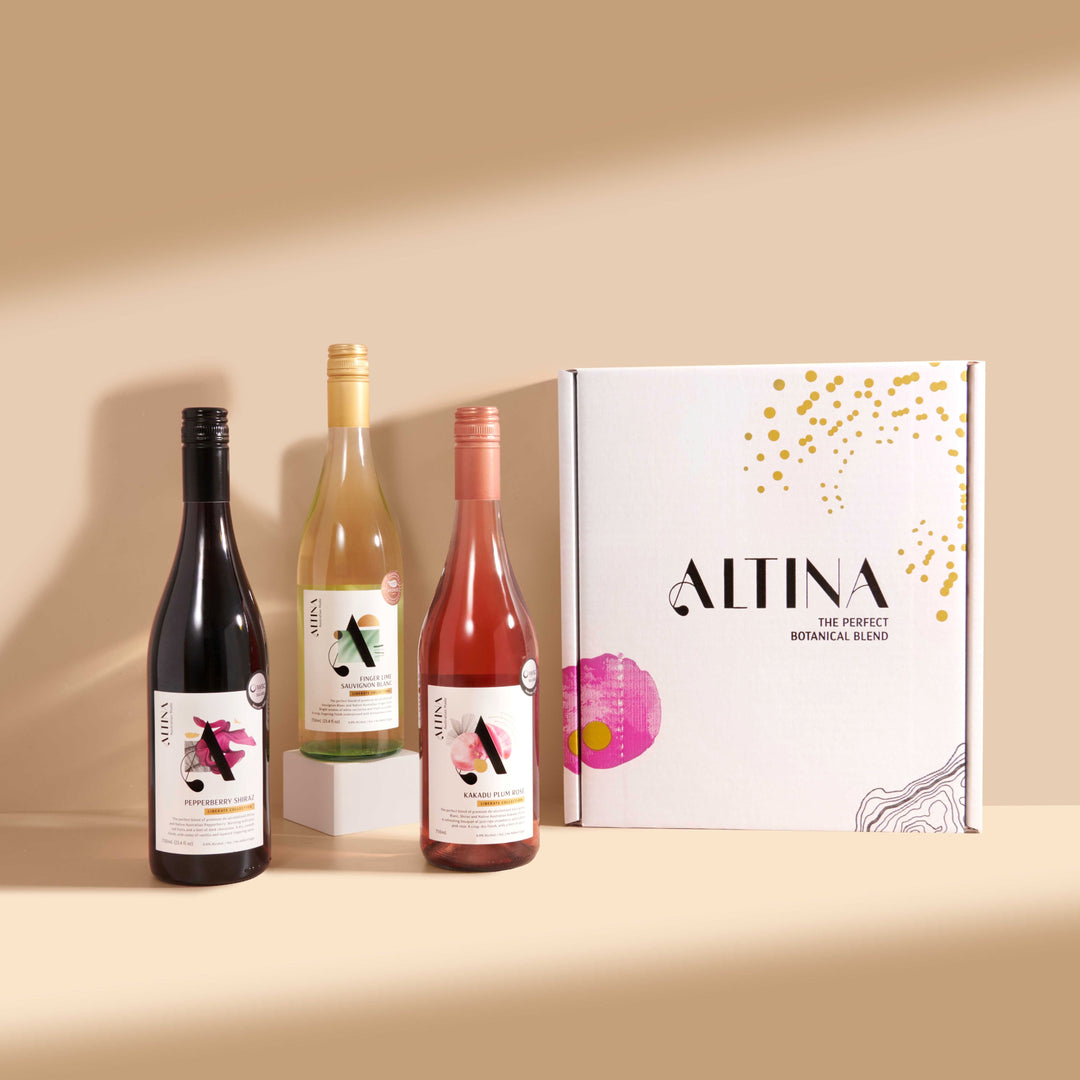 Three Altina still wine bottles arranged on a beige surface: a red wine on the left, a white wine elevated on a small rectangular block in the center, and a rosé wine on the right. A large white Altina gift box with botanical graphics stands to the right, set against a warm neutral background.