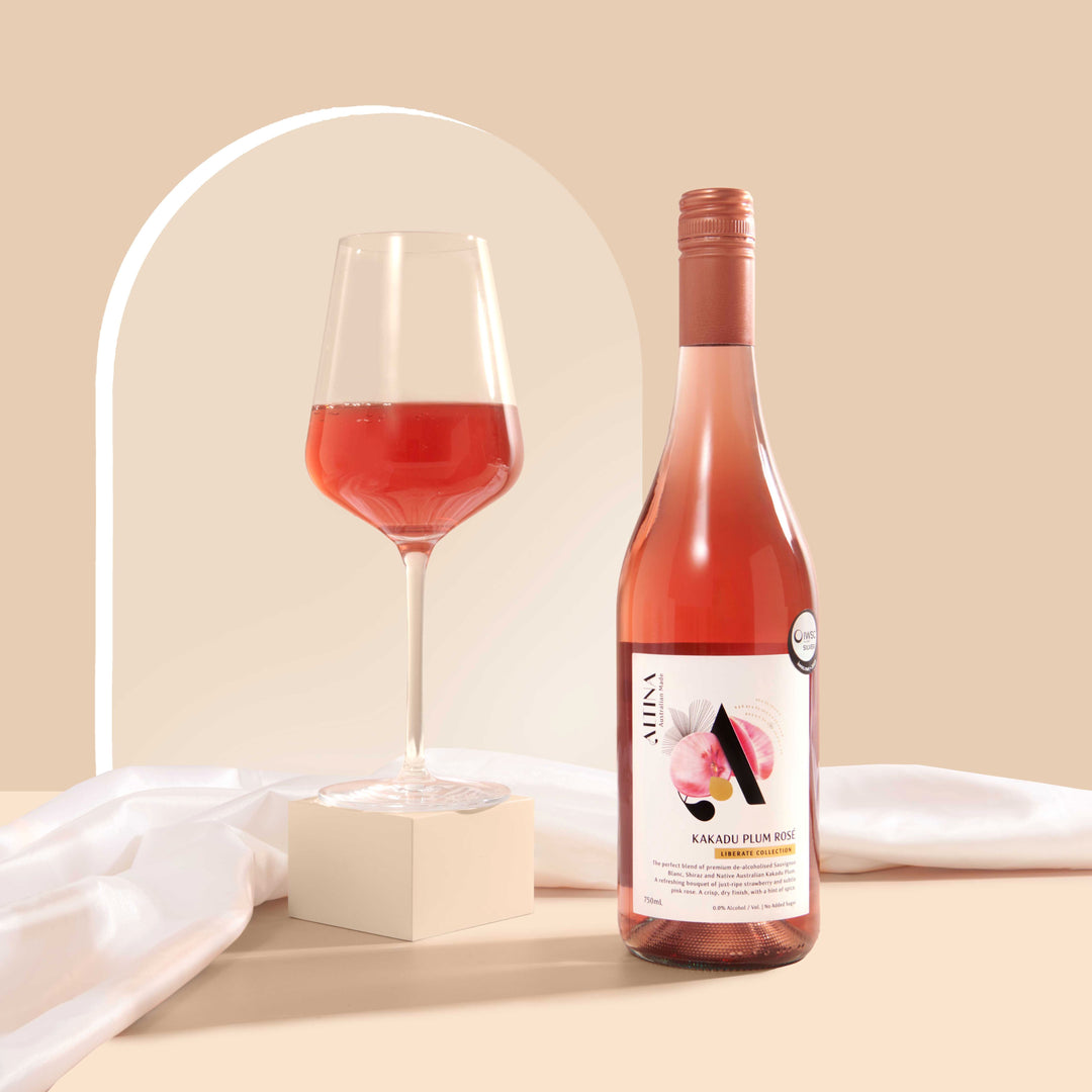 A glass of bright coral Kakadu Plum Rosé on a matte cream block beside an Altina Kakadu Plum Rosé bottle, standing on a beige surface with soft draped fabric and a subtle arched backdrop