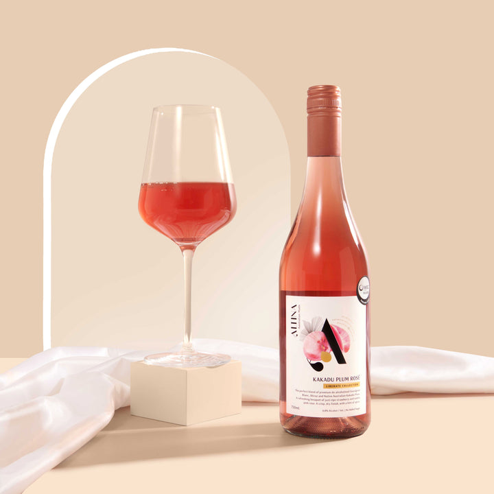 A glass of bright coral Kakadu Plum Rosé on a matte cream block beside an Altina Kakadu Plum Rosé bottle, standing on a beige surface with soft draped fabric and a subtle arched backdrop