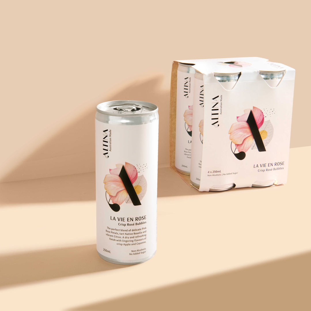 A single Altina non-alcoholic La Vie En Rosé slim can stands on a warm beige surface in the foreground, with a matching four-pack carton positioned behind it to the right.  Soft directional light creates long, clean shadows across the background.