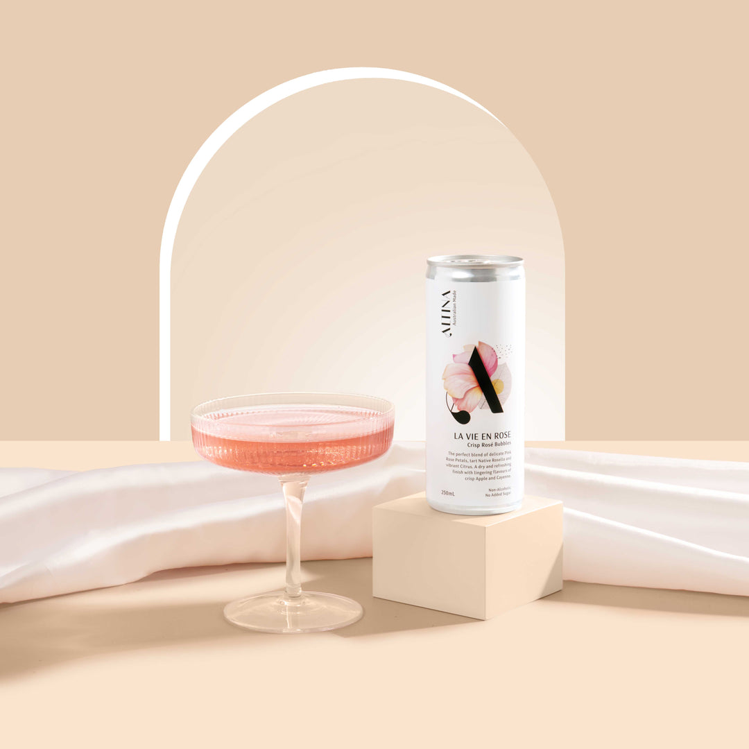 An Altina La Vie En Rose non-alcoholic canned rosé is displayed on a small plinth beside a shallow coupe glass filled with a pale pink drink, set against a soft beige background with a curved arch detail and draped fabric.