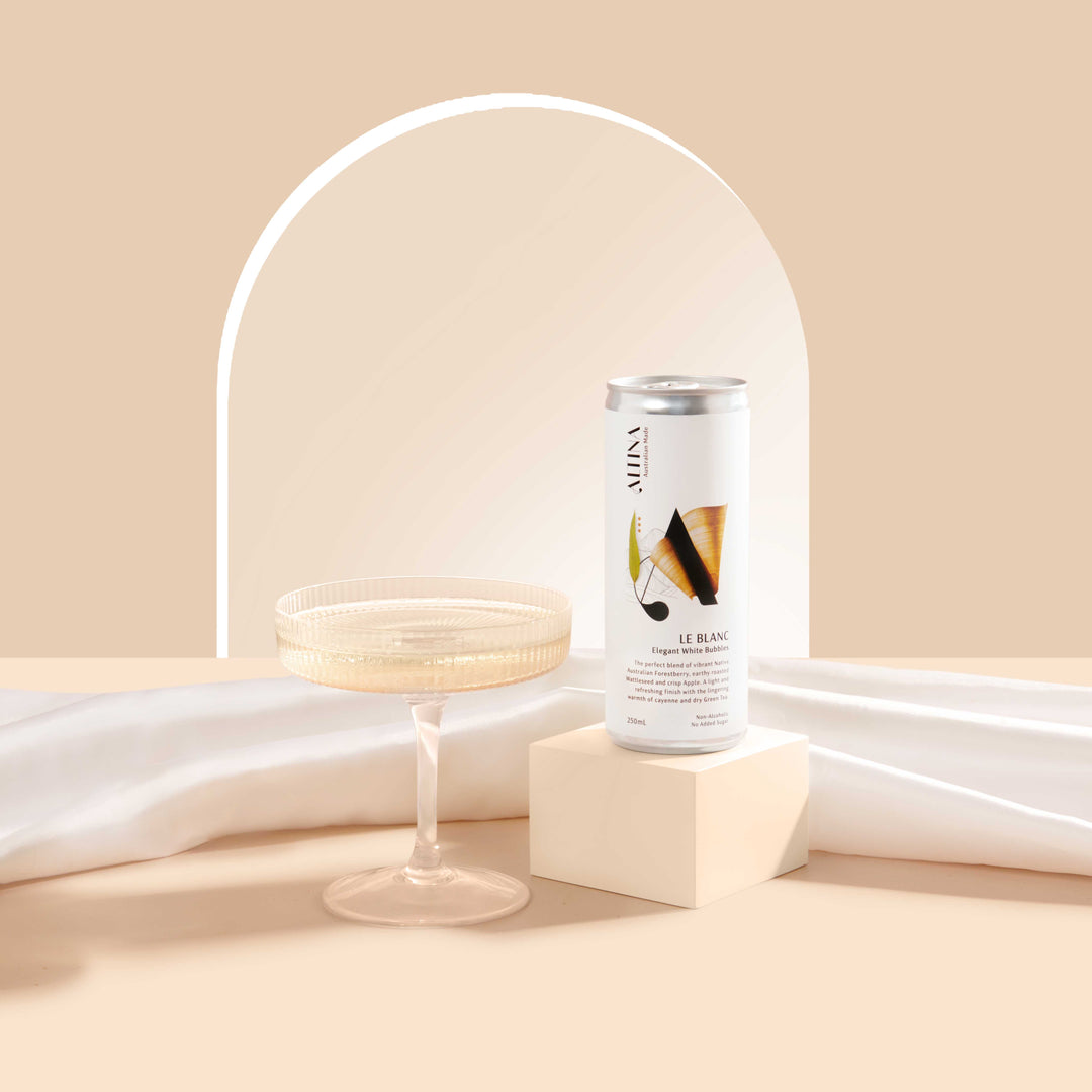 An Altina Le Blanc non-alcoholic canned white wine sits on a small plinth beside a shallow coupe glass, set against a soft beige background with a curved arch detail and draped white fabric.
