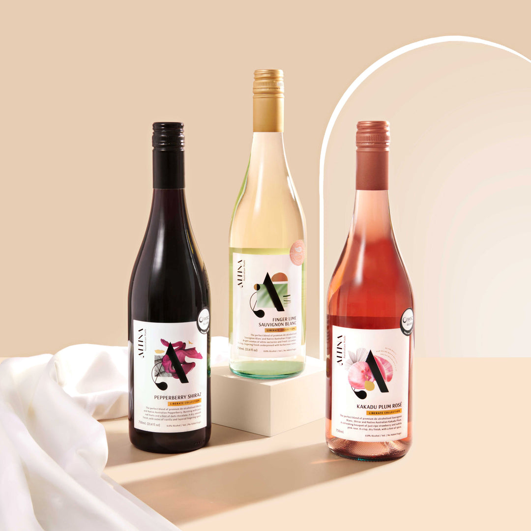 Three Altina Liberate still wines displayed on a beige surface: a white wine bottle elevated on a low rectangular block at the center, flanked by a red wine bottle on the left and a rosé wine bottle on the right. Set against a warm neutral background with a soft arched backdrop and draped white fabric
