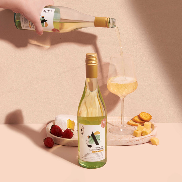 Altina de-alcoholised Finger Lime Sauvignon Blanc being poured into a wine glass, with a second non-alcoholic white wine bottle in the foreground beside a small plate of strawberries, cheese cubes, and crackers, set against a warm neutral background with soft shadows.