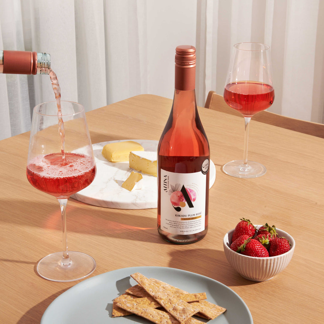 Altina de-alcoholised Kakadu Plum Rosé on a wooden table, with non-alcoholic rosé wine being poured into a wine glass. A bottle stands beside a second filled glass, with a marble plate holding sliced cheese in the background. In the foreground, a plate of breadsticks and a small bowl of strawberries sit on the table under soft natural light.