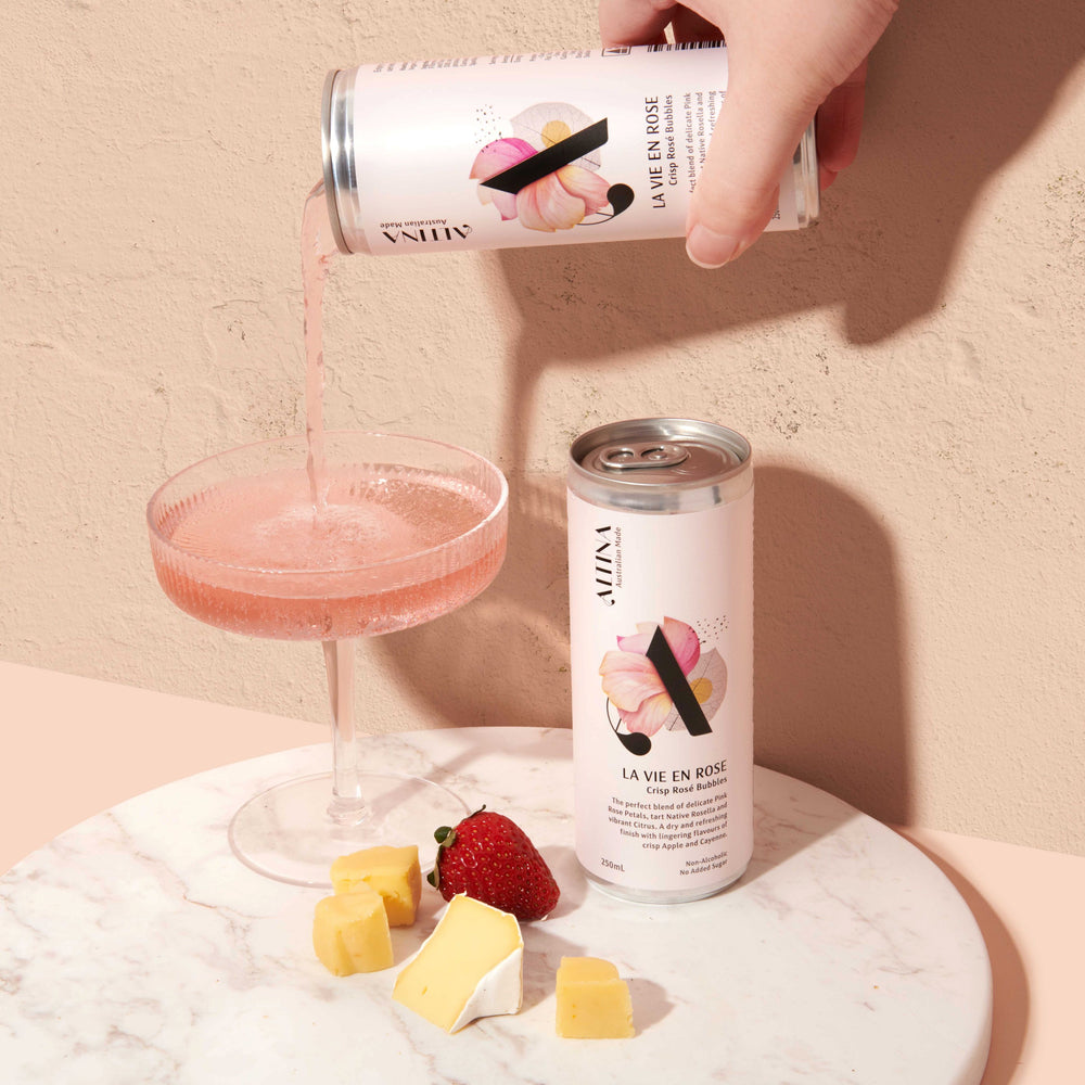 A hand pours pale pink de-alcoholised sparkling rose from an Altina La Vie En Rosé can into a shallow coupe glass. A second unopened non-alcoholic rose can stands beside the glass on a round marble surface. Small cubes of cheese and a strawberry are placed in front. The scene is set against a warm, textured beige background with soft, directional light.