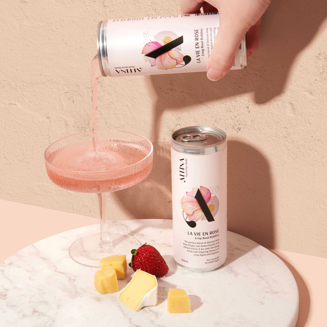 A hand pours pale pink de-alcoholised sparkling rose from an Altina La Vie En Rosé can into a shallow coupe glass. A second unopened non-alcoholic rose can stands beside the glass on a round marble surface. Small cubes of cheese and a strawberry are placed in front. The scene is set against a warm, textured beige background with soft, directional light.