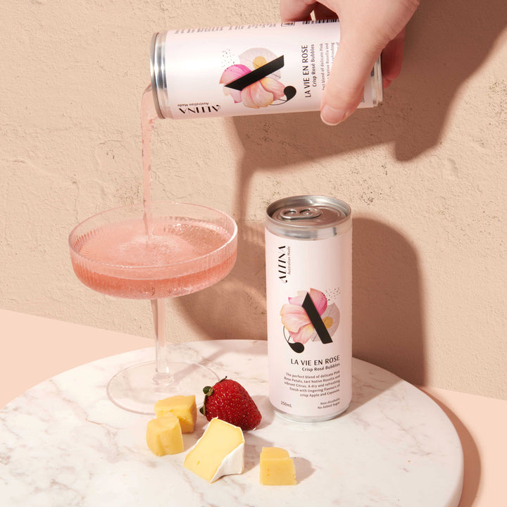 A hand pours pale pink de-alcoholised sparkling rose from an Altina La Vie En Rosé can into a shallow coupe glass. A second unopened non-alcoholic rose can stands beside the glass on a round marble surface. Small cubes of cheese and a strawberry are placed in front. The scene is set against a warm, textured beige background with soft, directional light.