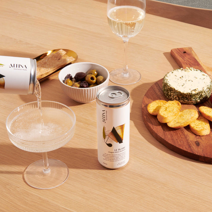 ALTINA LE BLANC is shown as a can pouring a pale non-alcoholic sparkling wine into a shallow coupe glass on a wooden table, with another can standing nearby. A wine glass with a light-coloured non-alcoholic white wine, a bowl of olives, and a wooden board with herb-coated cheese and crackers are arranged around it under warm natural light.