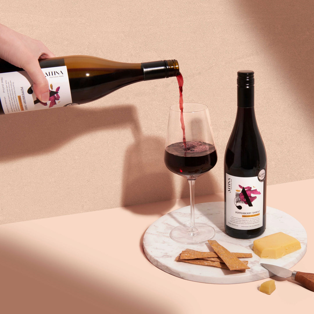 Altina Pepperberry Shiraz non-alcoholic red wine being poured into a clear stemmed glass on a pale surface. A bottle stands beside the glass on a round marble slab with rectangular crispbreads, a block of yellow cheese, and a small knife. Soft shadows fall across a textured neutral wall in warm light