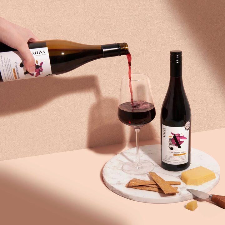 Altina Pepperberry Shiraz non-alcoholic red wine being poured into a clear stemmed glass on a pale surface. A bottle stands beside the glass on a round marble slab with rectangular crispbreads, a block of yellow cheese, and a small knife. Soft shadows fall across a textured neutral wall in warm light
