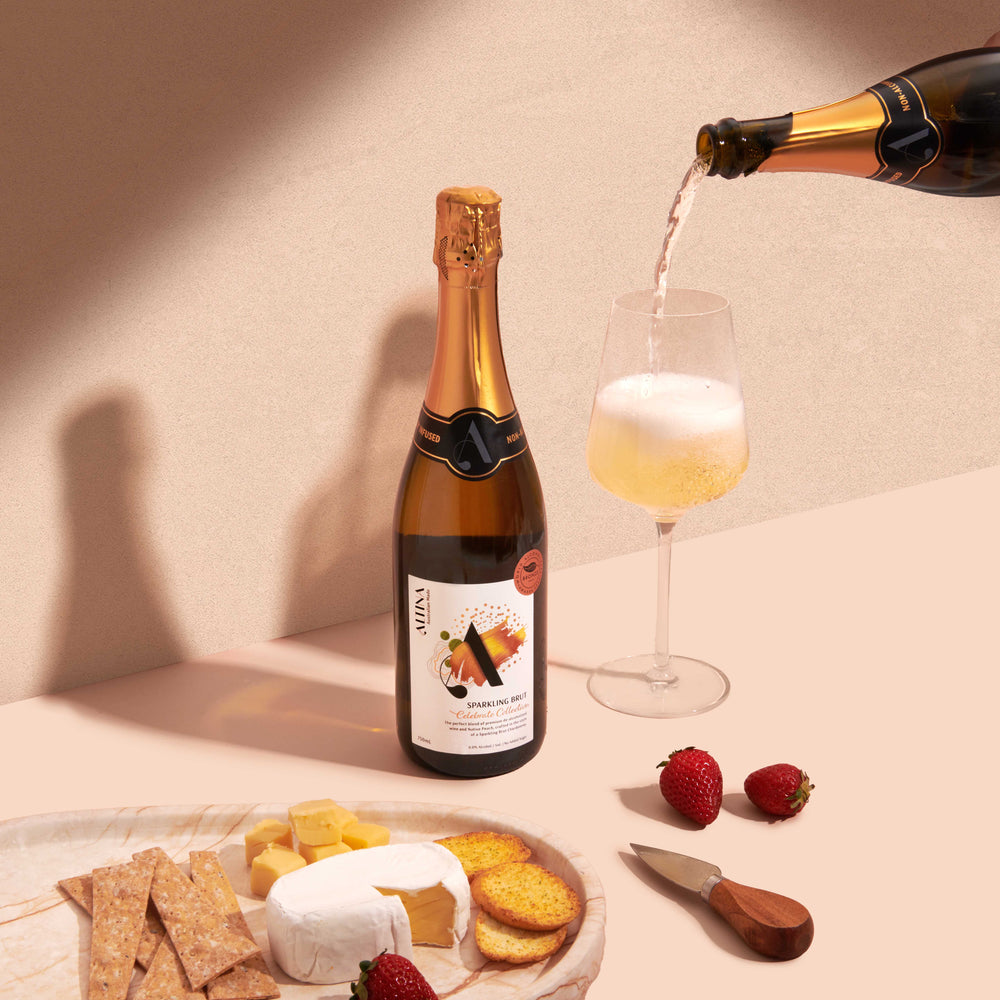 An Altina de-alcoholised Sparkling Brut bottle stands on a neutral surface as pale non-alcoholic sparkling wine is poured into a stemmed glass, forming a light foam. In the foreground, a stone plate holds crackers, cubed cheese, and a soft cheese wedge, with strawberries and a small cheese knife nearby, set against a warm textured background with soft angled shadows.