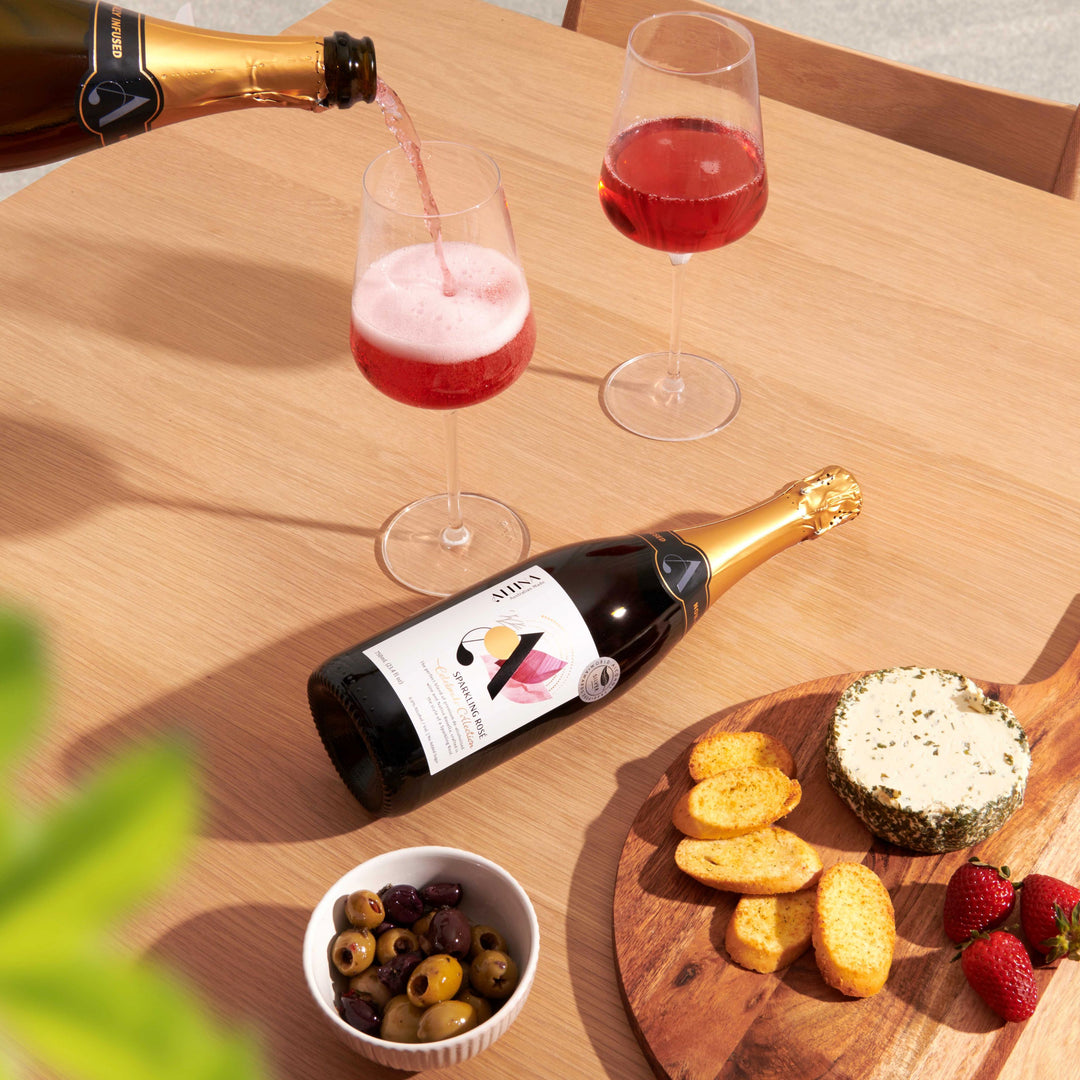 Altina non-alcoholic sparkling rosé is being poured from a bottle into a clear wine glass on a light wooden table, forming a soft pink foam. A second wine glass filled with rosé stands nearby. The bottle lies on its side with the label facing up. To the side, a wooden board holds a round herbed cheese, crackers, and strawberries, with a small bowl of mixed olives in the foreground. Soft natural light casts gentle shadows across the scene.