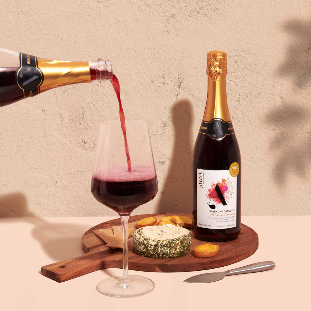 Altina de-alcoholised sparkling sansgria being poured into a wine glass, with a de-alcoholised sparkling red wine bottle beside a wooden board holding soft cheese and crackers, a small knife on the surface, and soft leaf shadows cast across a warm, textured neutral background.
