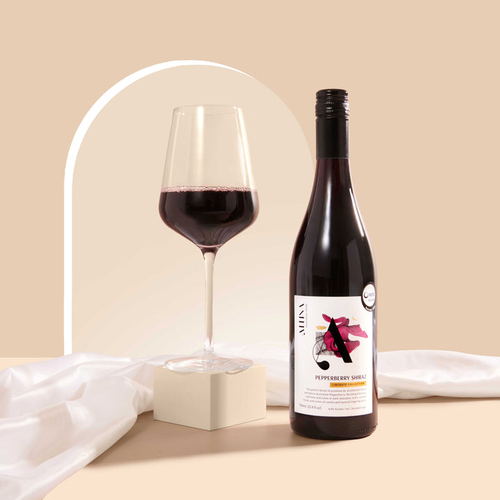 A glass of deep ruby Pepperberry Shiraz on a matte cream block beside an Altina Pepperberry Shiraz bottle, set on a beige surface with soft draped fabric and a subtle arched backdrop