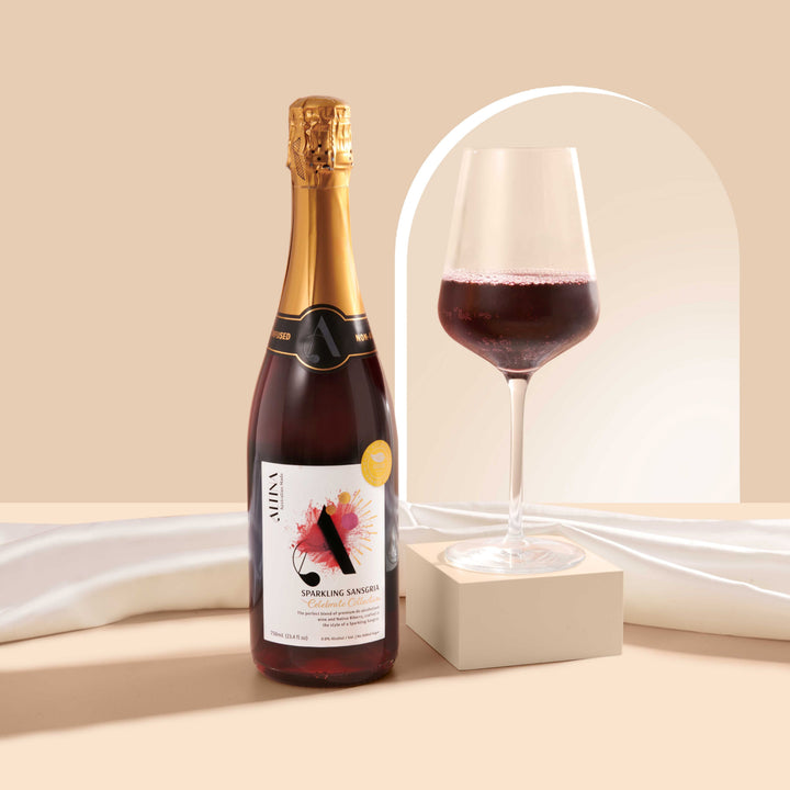 A glass of deep red sparkling sansgria on a cream rectangular block beside an Altina Sparkling Sansgria bottle, set on a beige surface with softly draped white fabric and a subtle arched backdrop.