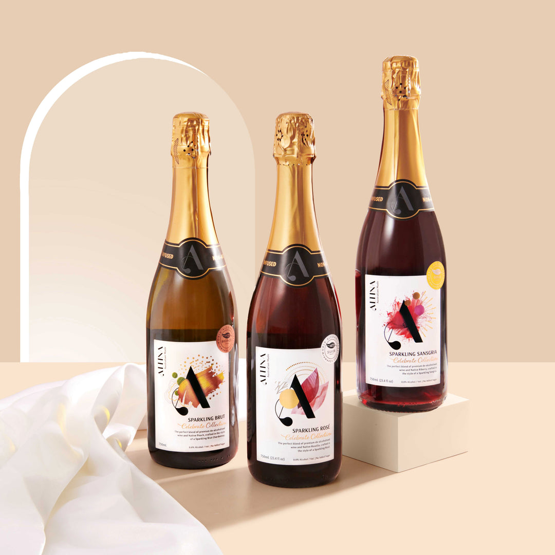 Three Altina sparkling wine bottles arranged on a beige surface, with one bottle elevated on a small rectangular block to the right and two bottles standing at the same level on the left. A soft white fabric drapes across the foreground, set against a warm neutral background with a curved light arch