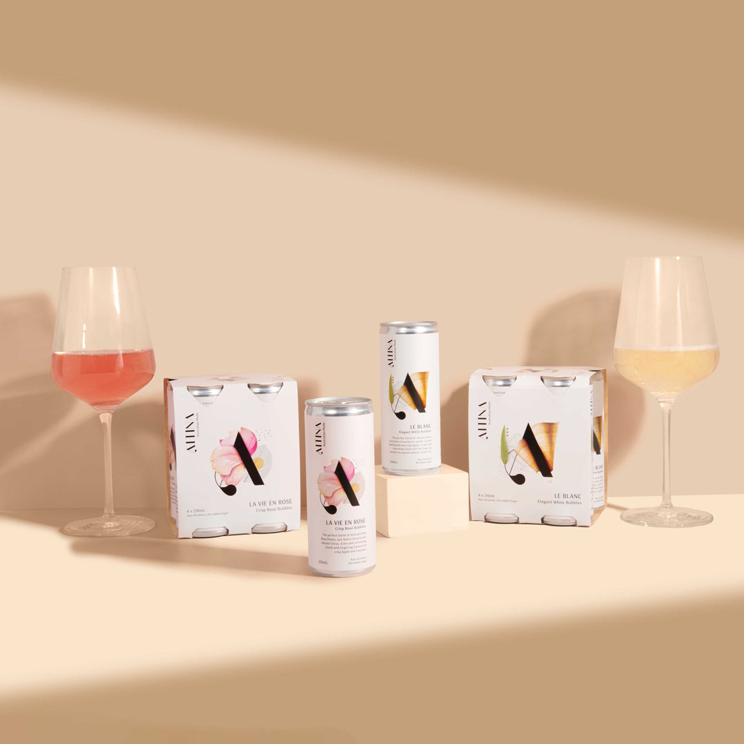 Altina wine cans with two four-pack boxes on a beige surface, a rosé glass on the left and a white wine glass on the right, one can raised on a small cream block and another can standing on the surface in front, set against a warm neutral background