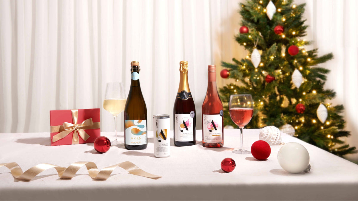 Three beverage bottles and a can arranged on a white tablecloth beside two wine glasses, a red gift box with a gold bow, red ornaments, ribbons, and a decorated Christmas tree in the background.